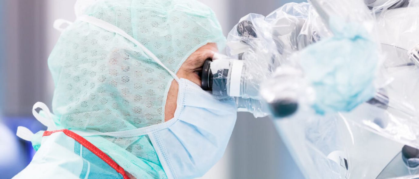 Ärztin mit Mundschutz und Schutzkleidung schaut während einer Operation durch ein Vergrößerungsokular