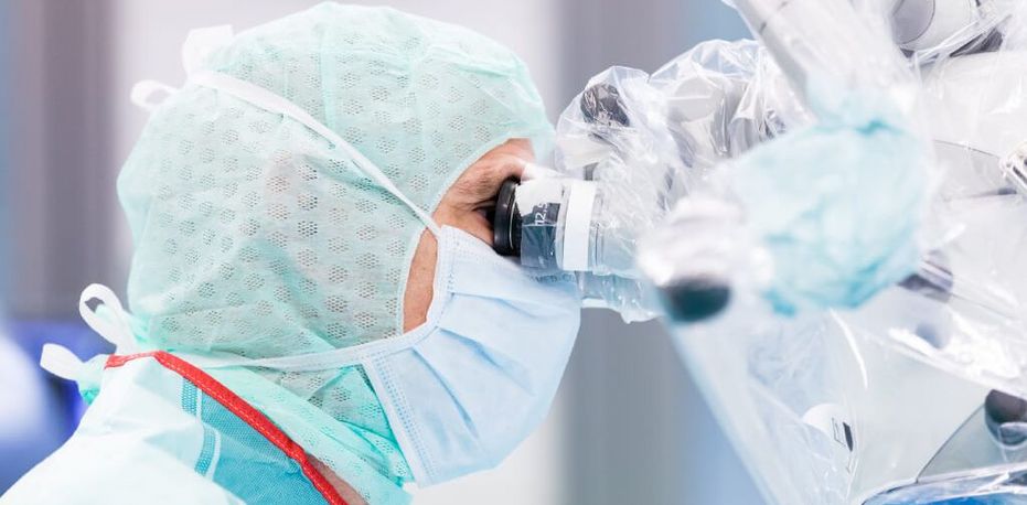 Ärztin mit Mundschutz und Schutzkleidung schaut während einer Operation durch ein Vergrößerungsokular